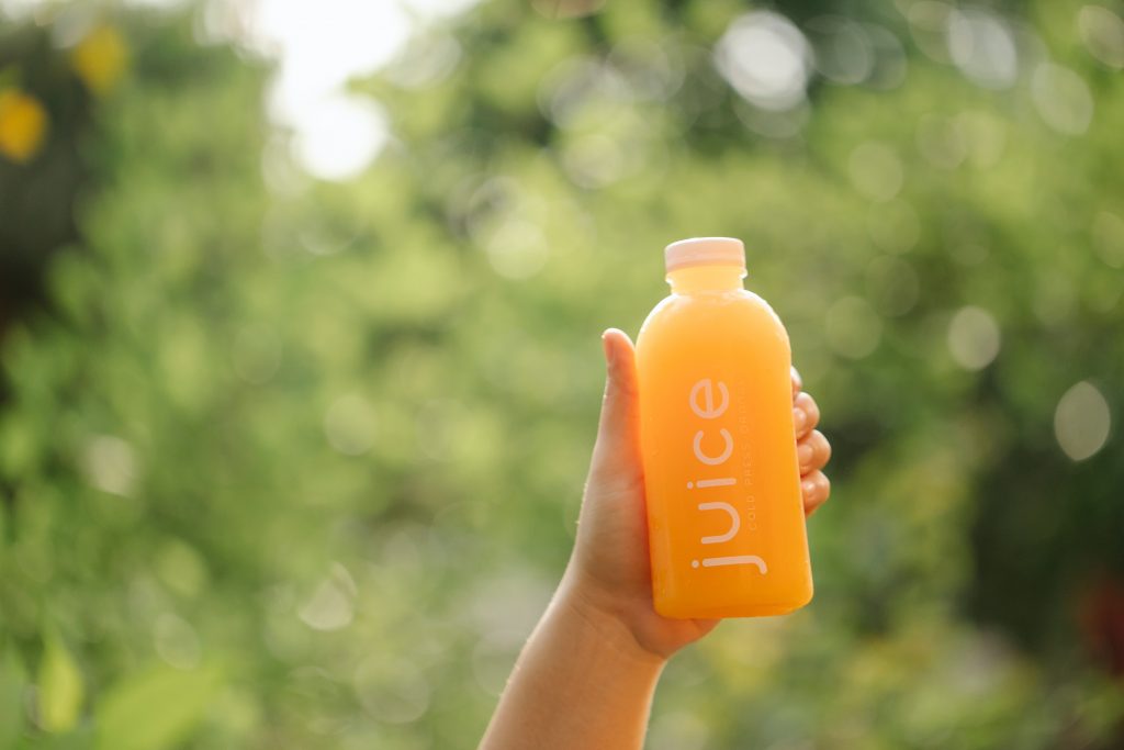 A hand holds a bottle of orange juice in front of a forest of trees